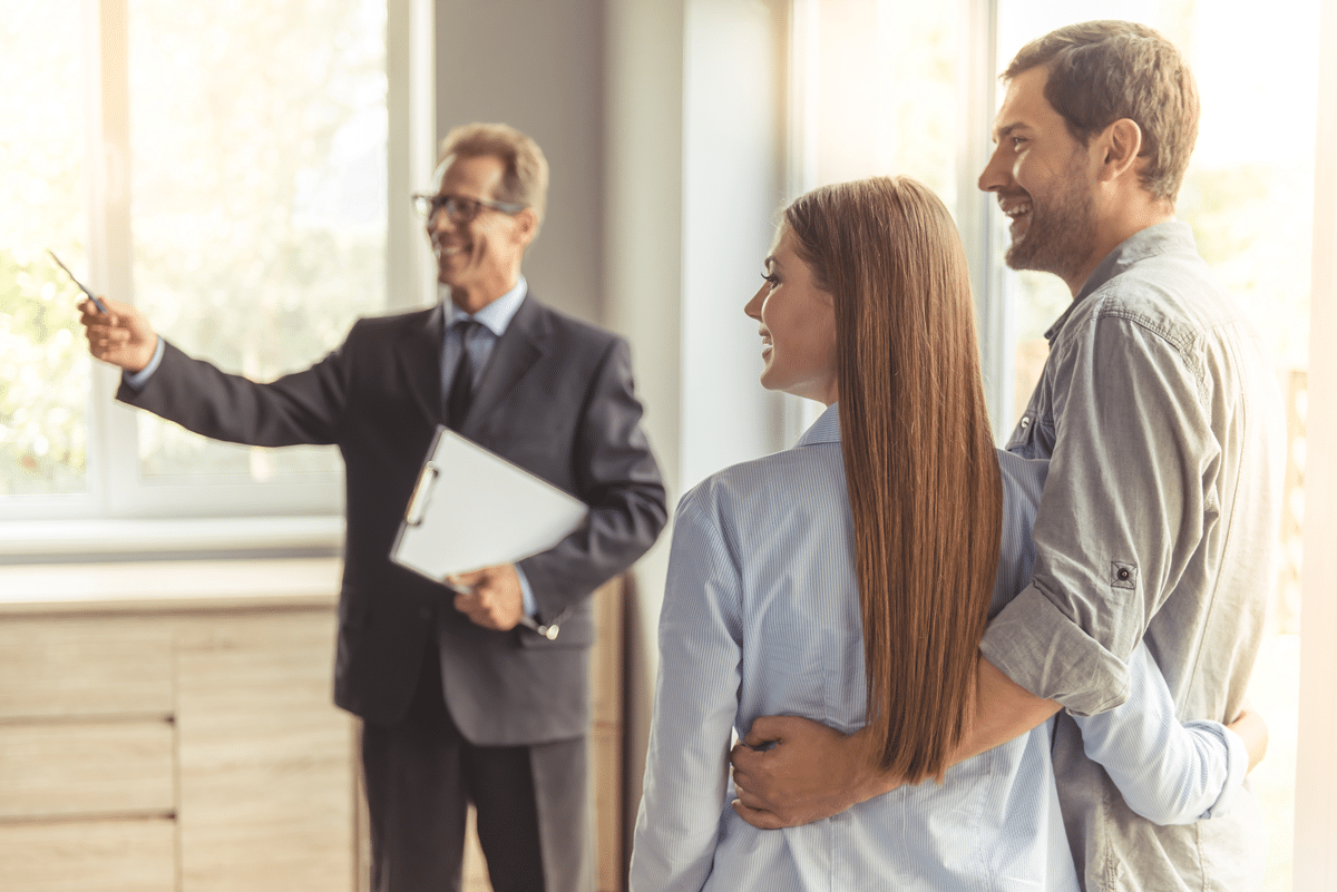 Un agent immobilier d'âge mûr, en costume classique et portant des lunettes, tient un carnet et sourit tout en faisant visiter un nouvel appartement à un jeune couple.